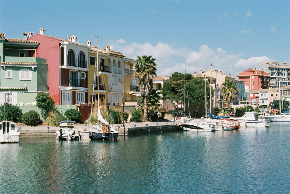 Brightly colored buildings line a serene marina with docked boats under a blue sky.